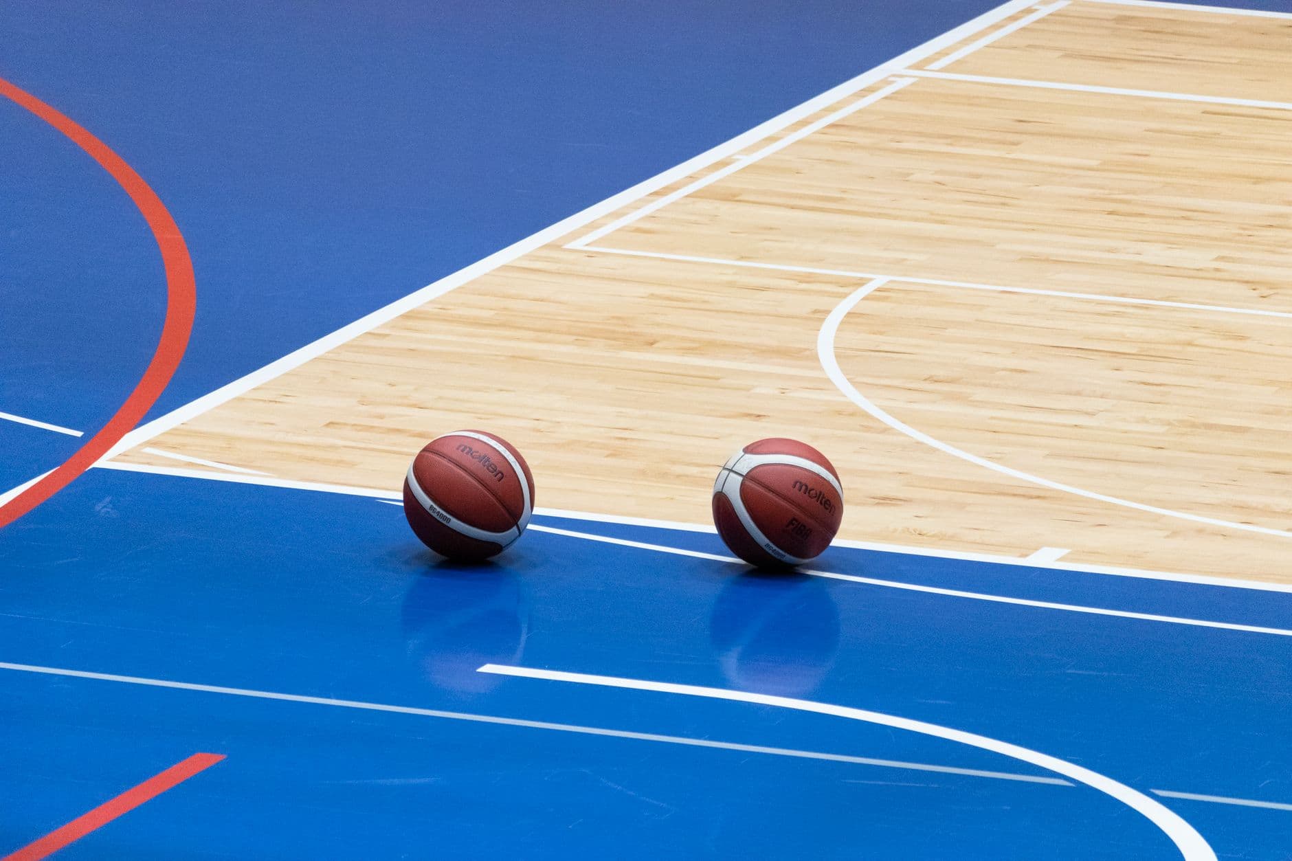 Two basketballs resting on a blue indoor basketball court with wooden flooring.